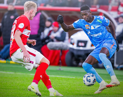 Два футболиста в действии на поле: один в красной форме, другой в синей, сражаются за мяч. Игра проходит на стадионе, в окружении зрителей. Видны элементы футбольной экипировки и игрового оборудования