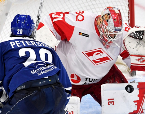 На изображении два хоккеиста у ворот. Игрок в синей форме атакует, а вратарь в белой и красной форме готовится отбить шайбу