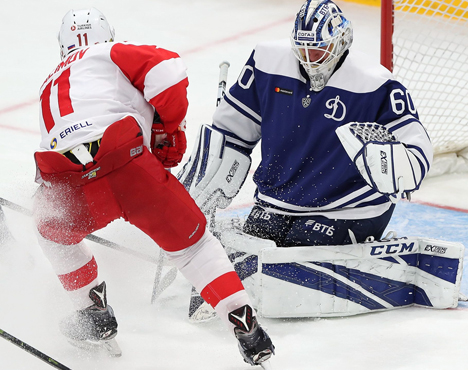 Хоккеист в красно-белой форме атакует ворота, защищаемые голкипером в синей форме. На фото запечатлен момент противостояния на льду. Вратарь готовится отразить удар шайбы