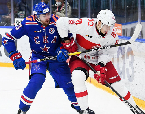 На изображении два хоккеиста в игровом моменте. Один в синей форме с буквой С на груди, другой в белой форме с красными элементами. Оба держат клюшки, соперничают за шайбу на льду